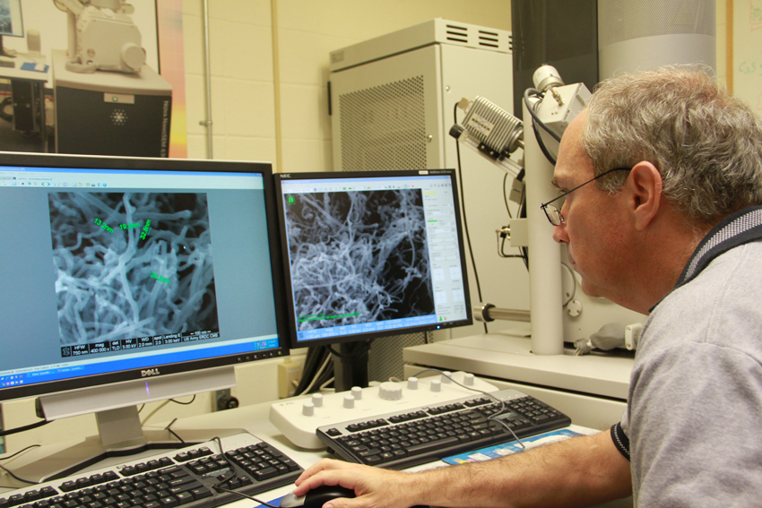 A man with grey hair wearing glasses sits at a desk in front of two monitors showing images of Scanning Electron Microscopy in a laboratory.
